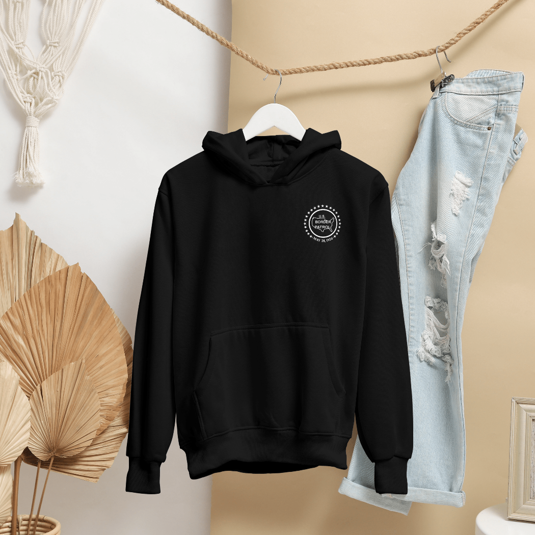 Black hoodie with a logo on a hanger next to light blue jeans against a neutral wall.