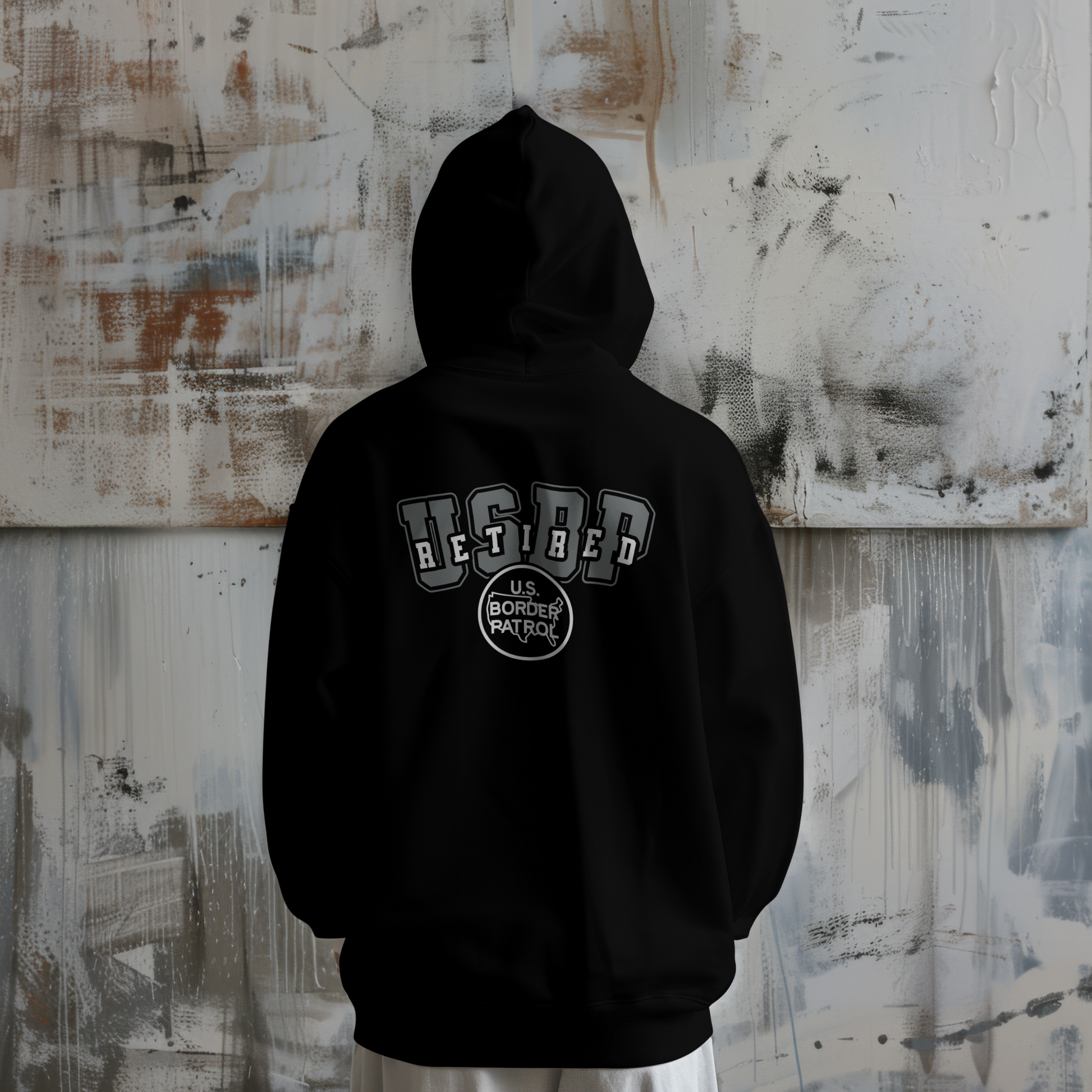 Black hoodie with 'Retired' and 'U.S. Border Patrol' text on the back, worn by a person against a textured wall.